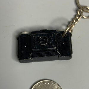 May include: Black camera keychain with a gold chain and a silver quarter for scale.