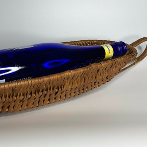 May include: A woven brown wicker basket with a blue glass bottle inside. The basket has two handles on the sides.