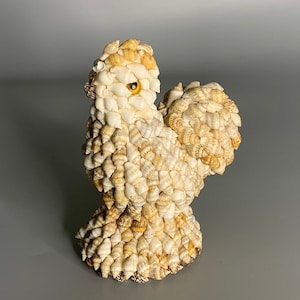 May include: A small, white, shell-covered sculpture of a rooster. The rooster has a yellow eye and is standing on a gray surface.