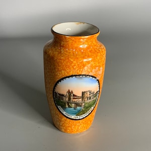 May include: A small orange ceramic vase with a white rim and a circular image of a bridge and buildings. The image is surrounded by a black border and the text "The Old Town Bridge, Salzburg, Austria".