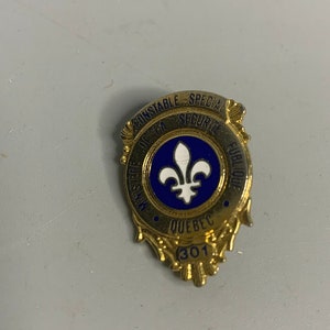 Puede incluir: Insignia de color dorado con esmalte azul. Dice "Constable Special Security Service Public Quebec" con una flor de lis en el centro y el número "301" en la parte inferior.