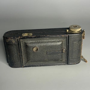 May include: A vintage folding camera in black with a leather exterior and brass accents. The camera is closed and has a lens cap on the front.