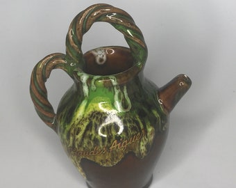 May include: A small brown ceramic pitcher with a green and brown glaze. The pitcher has a twisted rope handle and a spout. The words "Saudes Angeiras" are written on the side of the pitcher.