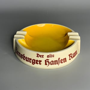 Puede incluir: Un cenicero de cerámica blanca con un interior amarillo y una inscripción roja que dice "Der alte Flensburger Hanfen Rum".