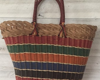 Puede incluir: Una bolsa de mano de mimbre tejido con un diseño a rayas en rojo, naranja, verde, azul y negro. La bolsa tiene dos asas de cuero marrón.