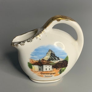 May include: A small white ceramic jug with a gold handle and trim. It features a painted image of a mountain range with a church and the text "Gornergrat Zermatt".