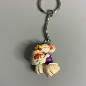 Könnte beinhalten: Ein Schlüsselanhänger mit einer weißen Comicfigur, die ein lilafarbenes Hemd trägt und ein gelbes und rotes Objekt in den Armen hält. Der Schlüsselanhänger ist an einem silbernen Metallring befestigt.