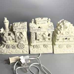 May include: Three off-white ceramic train cars with gold accents. The train cars have decorative details and are connected by a white electrical cord with small light bulbs. The train cars are a decorative item.