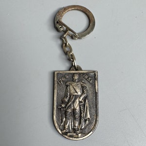 May include: A silver-toned keychain with a metal charm featuring a relief image of a person in a military uniform. The charm has the dates "1768" and "1858" inscribed on it.