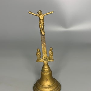 May include: A gold-toned metal bell with a figure of Jesus Christ on the cross on top. The bell has a round base and two figures on either side of the cross.