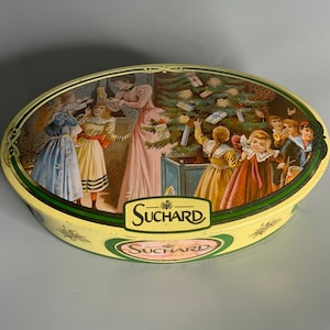 May include: Oval-shaped tin box with a Christmas scene featuring children around a decorated tree. The box is yellow with green trim and the text "SUCHARD" in pink lettering.