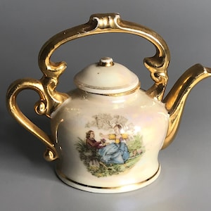 May include: A small, white porcelain teapot with a gold handle and spout. The teapot has a delicate floral design and a scene of two people in a garden.