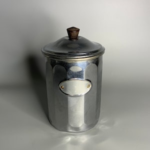 Könnte beinhalten: Eine silberne Metalldose mit einem Holzgriff auf dem Deckel. Die Dose hat ein kleines ovales Etikett mit goldener Umrandung.