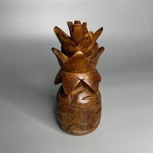 Puede incluir: Una escultura de piña de madera tallada. La piña es de color marrón cálido con tallas detalladas de hojas y la corona de la piña. La escultura está de pie sobre un fondo gris liso.