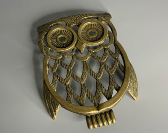 May include: A brass owl-shaped trivet with a cutout design. The owl has large eyes and a detailed feather pattern.