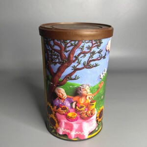 May include: A cylindrical tin with a brown lid and a colorful illustration. The artwork depicts two people at a picnic table with food, surrounded by sunflowers, a tree, and a blue sky.