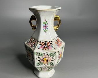 May include: White ceramic vase with gold accents and floral designs. The vase has a hexagonal shape and two gold handles. The floral designs are in shades of pink, red, green, and yellow.