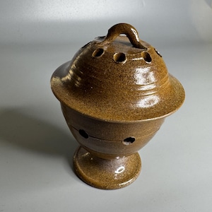 May include: A brown ceramic incense burner with a lid and handle. The burner has a round base and a bowl-shaped body with several small holes for ventilation. The lid also has holes. The ceramic has a speckled, glazed finish.