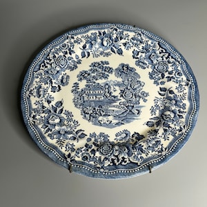 May include: A blue and white ceramic plate with a floral pattern and a central image of a house and a bridge. The plate has a scalloped edge and a beaded border.