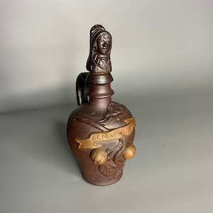 May include: A brown ceramic bottle with a sculpted design of a woman's head on the stopper and a banner that reads "Calvados".