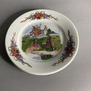 Puede incluir: Un plato de cerámica blanco con un borde floral y una escena pintada de dos personas caminando junto a una iglesia. La escena se desarrolla en un campo verde con un pequeño puente de madera.