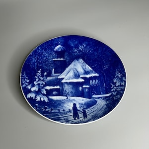 Puede incluir: Un plato de cerámica azul y blanco con una escena de invierno. El plato presenta una iglesia, una casa y dos figuras caminando en la nieve. El plato está decorado con un esmalte azul y blanco.