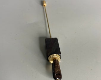 Puede incluir: Un alfiler de madera y oro con un mango de madera marrón oscuro y un alfiler de metal dorado. El mango tiene un patrón moteado.