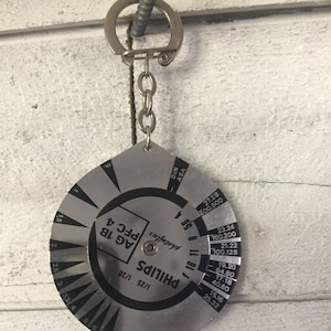 May include: A silver-colored metal keychain with a circular dial featuring a Phillips screwdriver size chart. The dial has markings for different sizes of Phillips screwdrivers, along with corresponding DIN and ASA values. The keychain also includes a small metal tag with the words "shotoflux" and "PFC 4" printed on it.