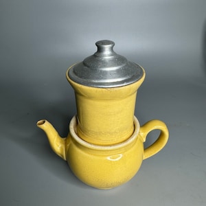 Op de afbeelding: Een gele theepot van keramiek met een zilveren deksel. De theepot heeft een rond lichaam en een tuit aan de zijkant. Het deksel is van metaal en heeft een afgeronde bovenkant.