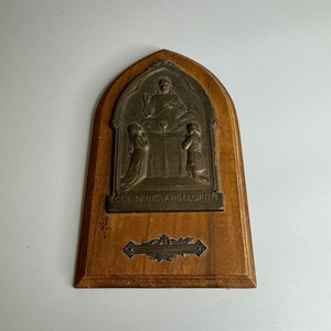 Puede incluir: Una placa de madera con un relieve plateado de Jesús partiendo el pan con dos figuras arrodilladas. La placa tiene la inscripción "Ecce Panis Angelorum" y un diseño floral decorativo.