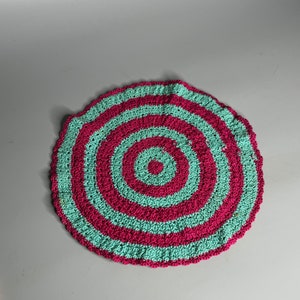 May include: A round crocheted coaster with alternating pink and teal stripes. The center of the coaster has a smaller circle of teal surrounded by a pink circle.