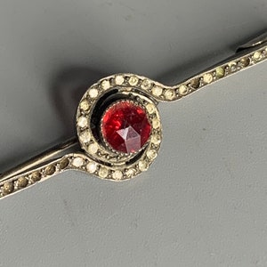 Puede incluir: Un broche de plata con una piedra roja rodeada de piedras blancas más pequeñas. El broche tiene forma de espiral.