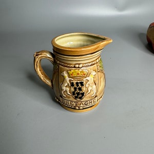 May include: A brown ceramic pitcher with a handle and a design of a coat of arms with two horses and the word "BRETAGNE" on the front.