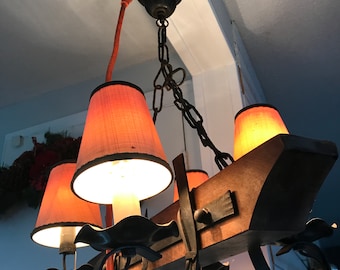 May include: A rustic chandelier with four orange lampshades. The fixture features a wooden beam, decorative metal accents, and a chain for hanging. The lampshades are a warm orange color, and the light bulbs are lit.