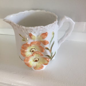 May include: A white porcelain pitcher with a floral design. The pitcher has a scalloped rim and a handle. The flowers are orange, yellow, and green.