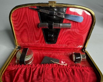 May include: A black leather travel case with a gold trim and a red velvet interior. The case contains a small silver container, a black and silver container, a silver tweezers, and a silver nail file.