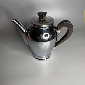May include: A chrome-plated teapot with a dark brown handle and lid knob. The teapot has a rounded body, a spout, and a base. The handle is curved and attached to the side of the teapot. The lid knob is square. The teapot is on a white surface.