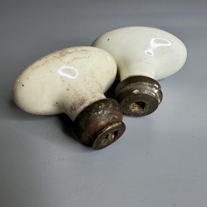 May include: Two antique white porcelain door knobs with aged brass bases. The oval-shaped knobs show signs of wear and age, with slight discoloration and patina on the metal. These vintage knobs are a classic hardware element.