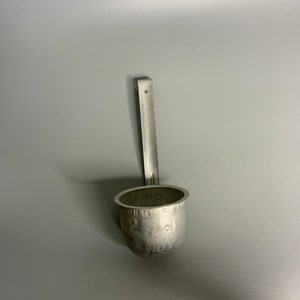 May include: A silver metal ladle with a long handle. The ladle has markings that read "2 oz. of the liquid".