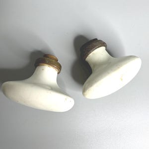 Puede incluir: Dos pomos de puerta antiguos de porcelana blanca con detalles metálicos color latón. Los pomos tienen forma ovalada y redondeada y están diseñados para un agarre fácil. Estos pomos vintage son ideales para añadir un toque de elegancia clásica a cualquier puerta.