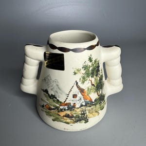 Może przedstawiać: Kremowy ceramiczny kubek z dwoma uchwytami, z ręcznie malowaną sceną krajobrazu z chatą, drzewami i górami. Obrzeże zdobi powtarzający się brązowy wzór. Mała etykieta jest przymocowana z boku.