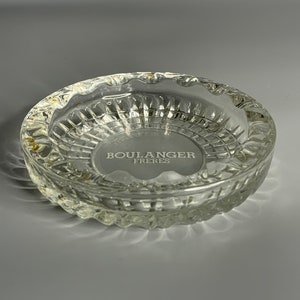 Pode incluir: Um cinzeiro de vidro transparente com uma borda texturizada elevada e uma inscrição circular que diz "BOULANGER FRERES".