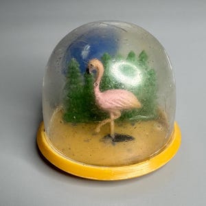 May include: A pink flamingo figurine inside a clear glass dome with a yellow base. The dome is filled with green and blue plastic trees.