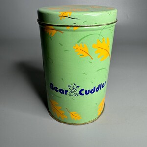 May include: A cylindrical green tin with a lid, decorated with yellow leaf designs and the words "Bear Cuddle" in blue. The tin is a light green color.