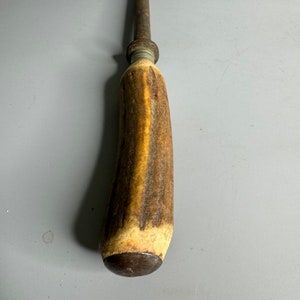 May include: A vintage steel knife sharpener with a brown antler handle. The handle has a dark brown stain and a metal ring at the top.