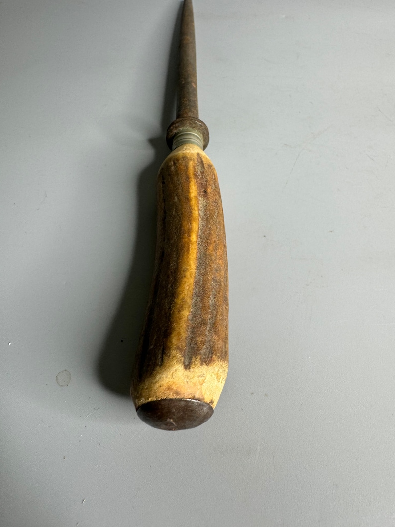 May include: A vintage steel knife sharpener with a brown antler handle. The handle has a dark brown stain and a metal ring at the top.