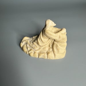 Könnte beinhalten: Eine weiße Keramikskulptur einer Person, die im Gebet kniet und die Hände gefaltet hat. Die Skulptur ist detailliert mit fließenden Roben und langen, fließenden Haaren.