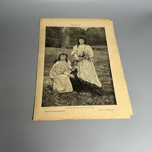 May include: A black and white photograph of two girls in dresses standing in a grassy field. The girl on the left is holding a black dog. The girl on the right is holding a bouquet of flowers. The text "DANS LA PRAIRIE.  D'après J.-Léon Gérôme" is printed below the image.