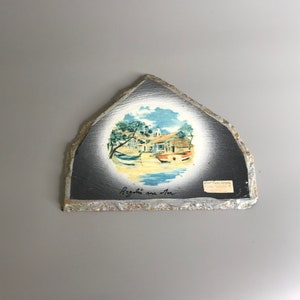 Könnte beinhalten: Ein schwarzer Schieferuntersetzer mit einem gemalten Bild einer kleinen Stadt mit Booten im Wasser. Der Text "Regalo sur mer" ist auf dem Untersetzer geschrieben.