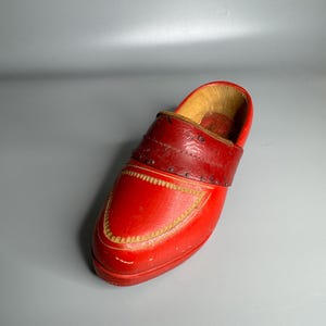 May include: A single, bright red wooden clog with a dark red leather strap across the top. The toe of the clog has a decorative stitched design in a lighter color. The interior is a light brown color.